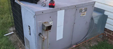 An all-in-one packaged HVAC unit rests beside a brick home, mid-service with a technician's tool still resting on top.